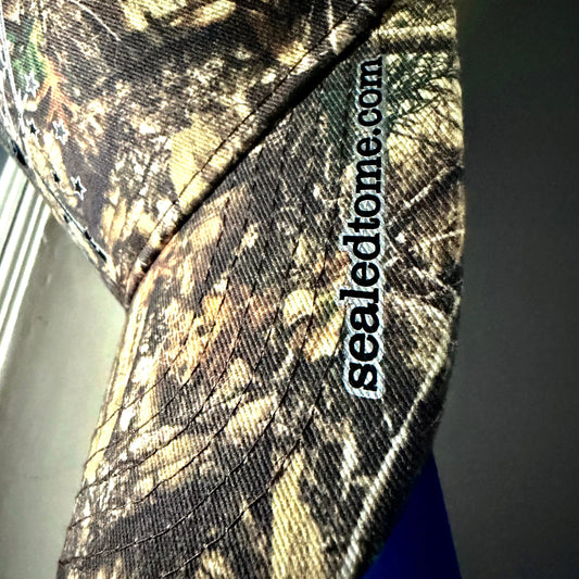 Close-up of a camouflage baseball cap with a visible 'sealedhome.com' embroidered patch on the side, showing a brown, beige, and black woodland camo pattern.