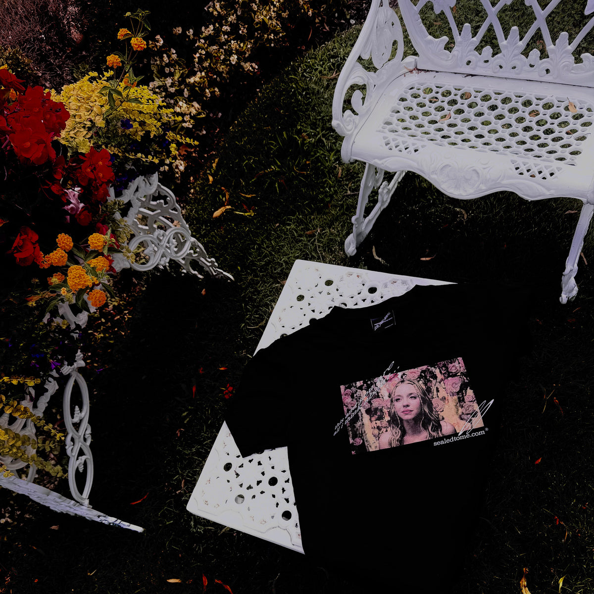A black knit garment laid flat on dark grass, surrounded by white decorative bench pieces and a small printed photo card. No person or animal is present, but the outdoor garden-like setting makes this a Lifestyle image rather than a clean studio Product/Packaging shot.