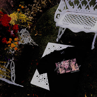 A black knit garment laid flat on dark grass, surrounded by white decorative bench pieces and a small printed photo card. No person or animal is present, but the outdoor garden-like setting makes this a Lifestyle image rather than a clean studio Product/Packaging shot.