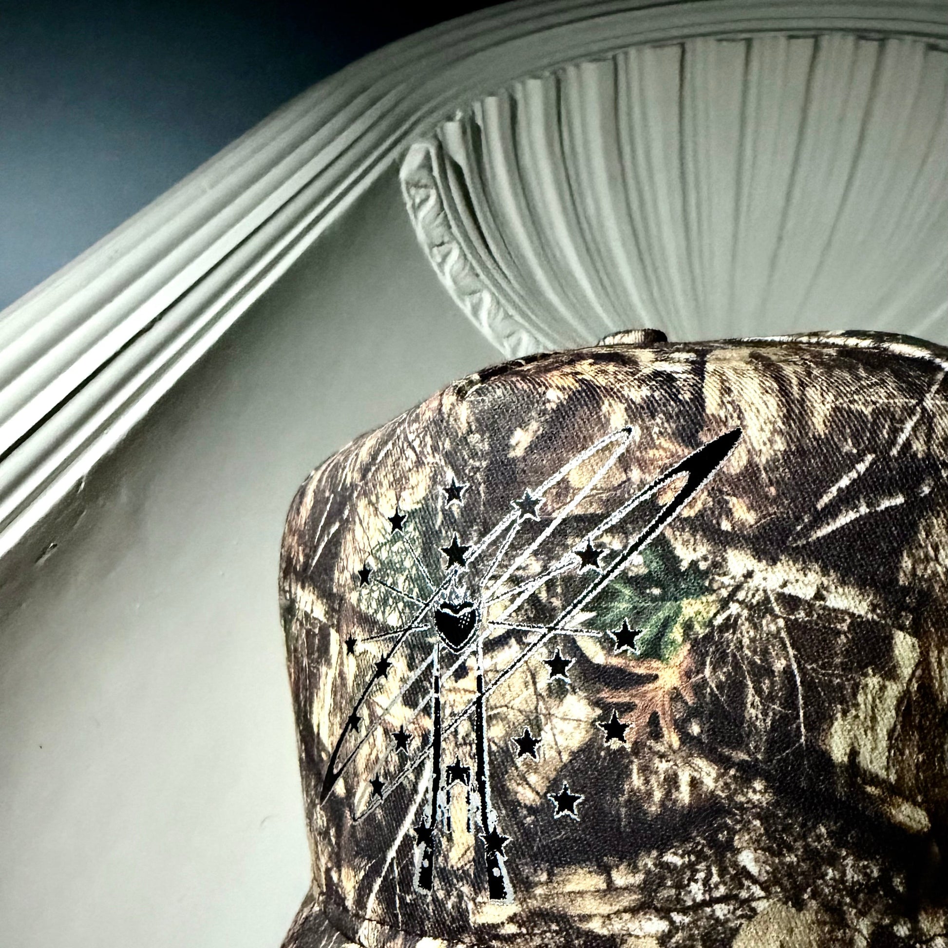 A close-up of a camouflage baseball cap featuring a black and silver graphic of a cowboy hat with stars and a bowtie on the front panel.