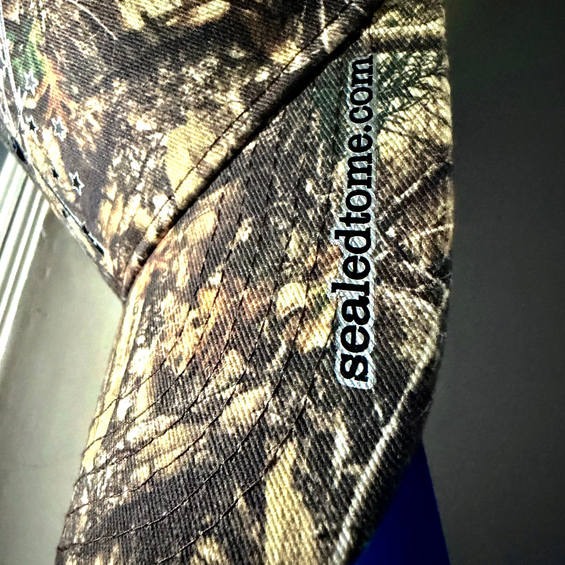Close-up of a camouflage baseball cap with a visible 'sealedhome.com' embroidered patch on the side, showing a brown, beige, and black woodland camo pattern.