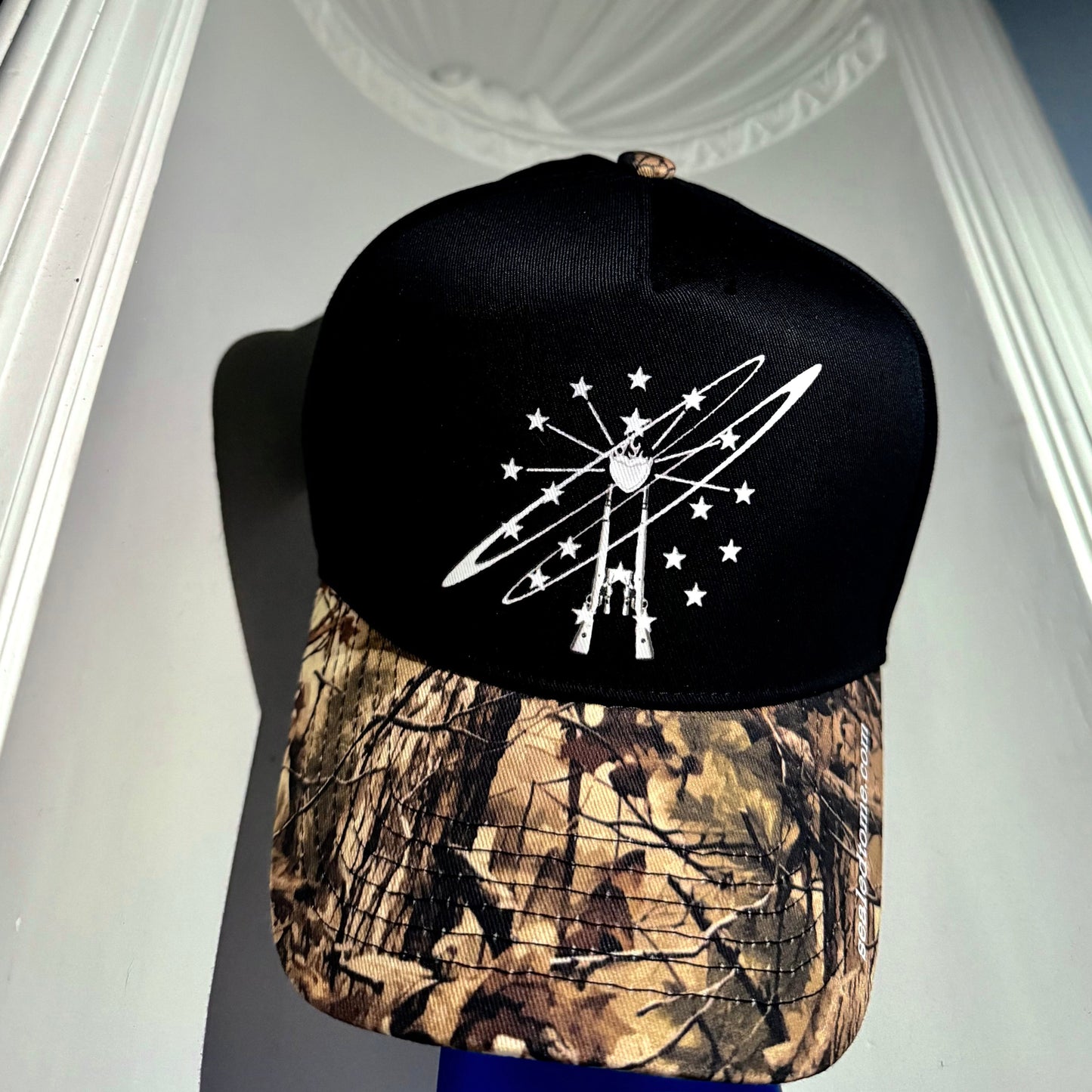 A black snapback cap with a white graphic design on the front and a brown camouflage patterned brim, displayed on a blue stand against a white architectural background.