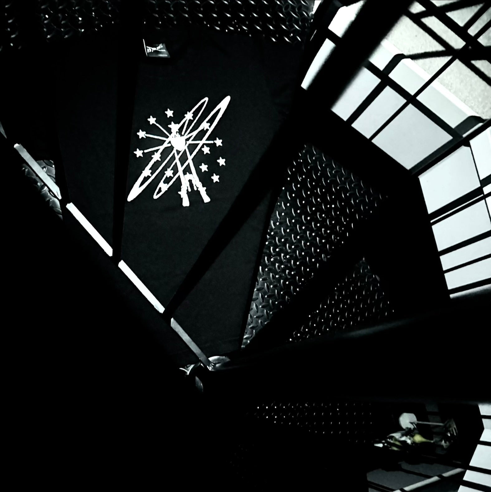 A dramatic high-contrast black and white photograph of a spiral staircase viewed from below, featuring a white graphic logo of a stylized figure with stars on the black handrail.